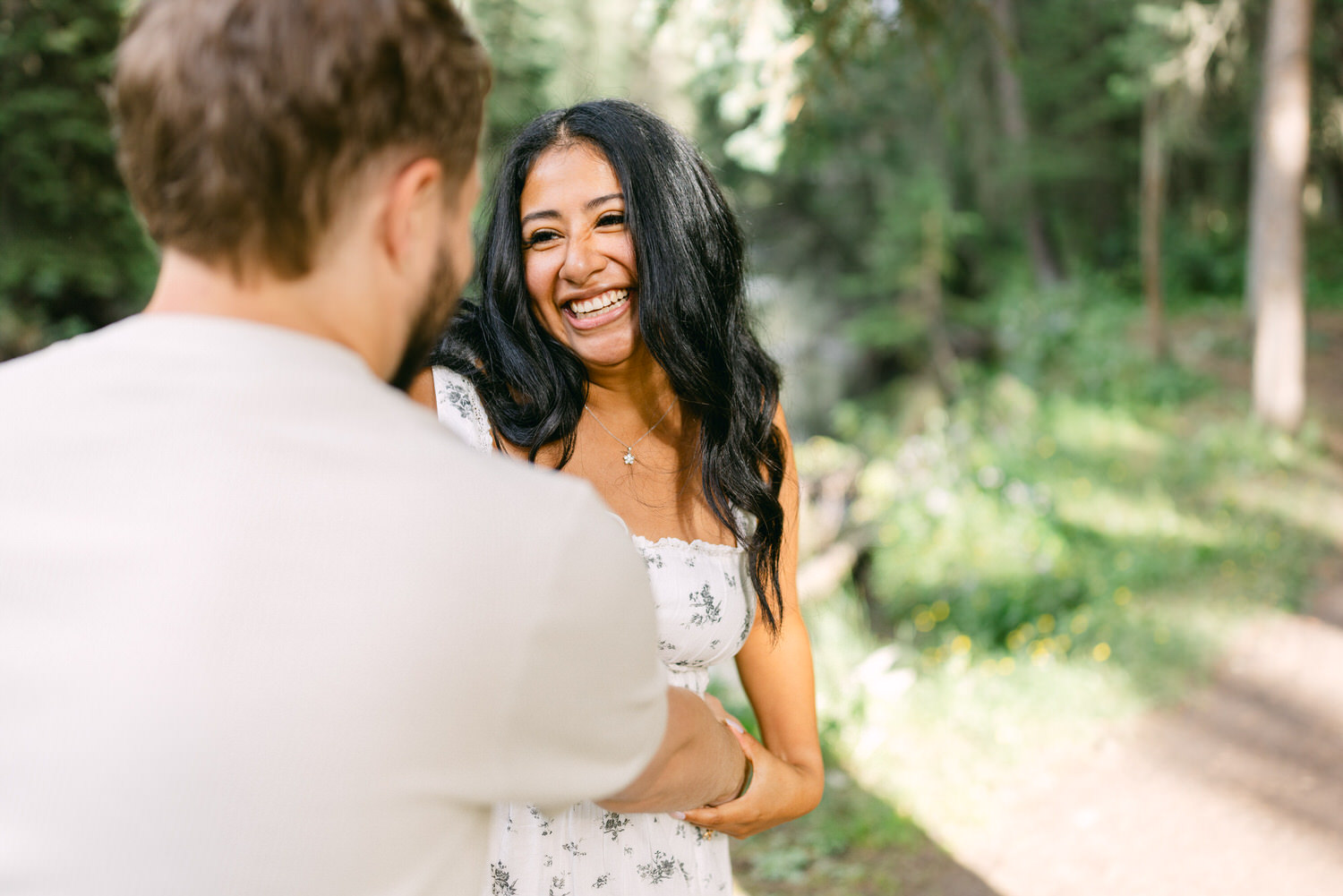 Woman with a big smile holding hands with a man in a sunny forest setting.