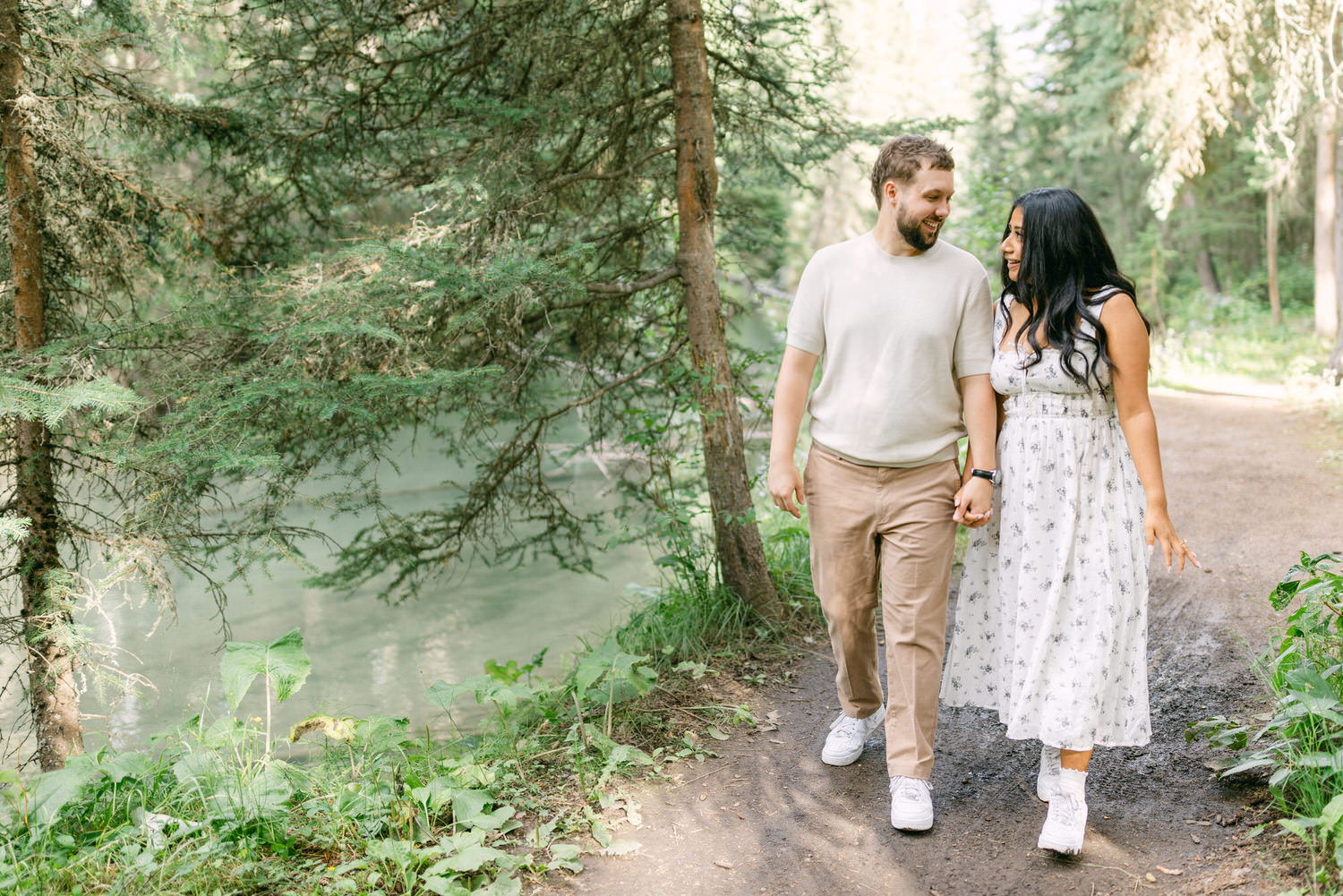 A man and a woman walking together holding hands on a forest path, with lush greenery around and a small stream beside them.