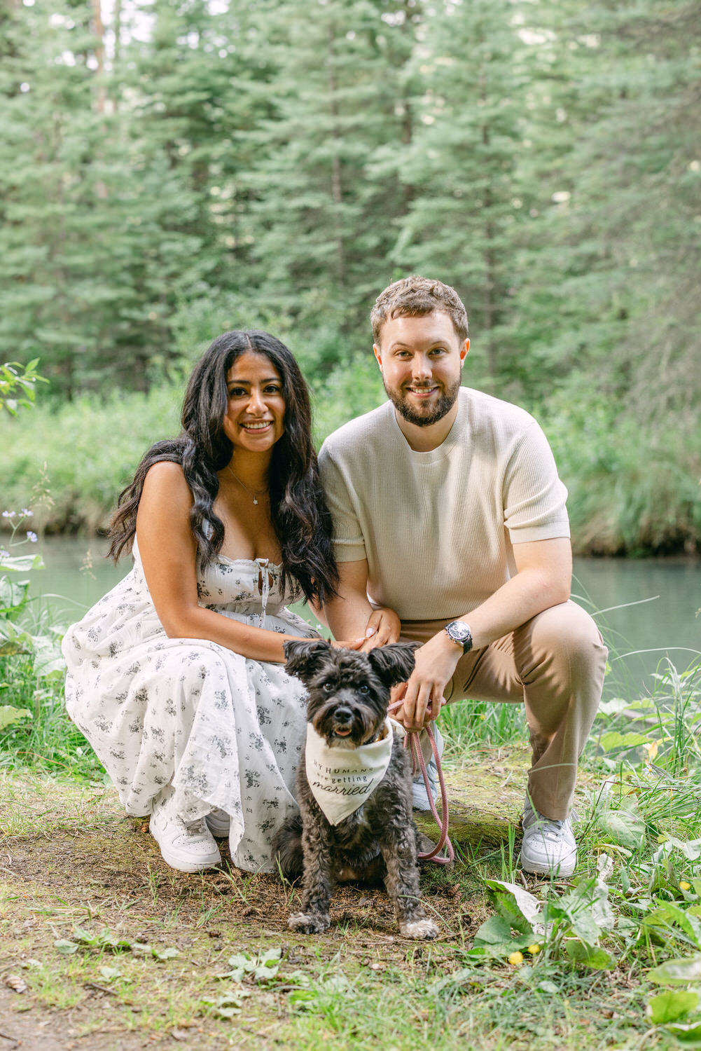 A smiling couple and their dog sitting by a forest stream, with the dog wearing a "My humans are getting married" bandana.