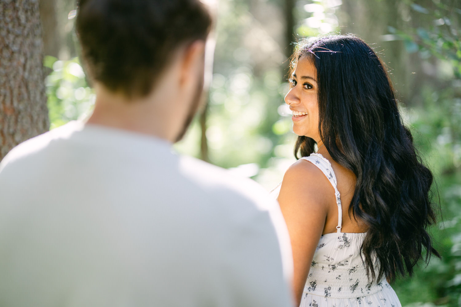 Woman in a white dress smiling and looking over her shoulder at a man in a forest setting.