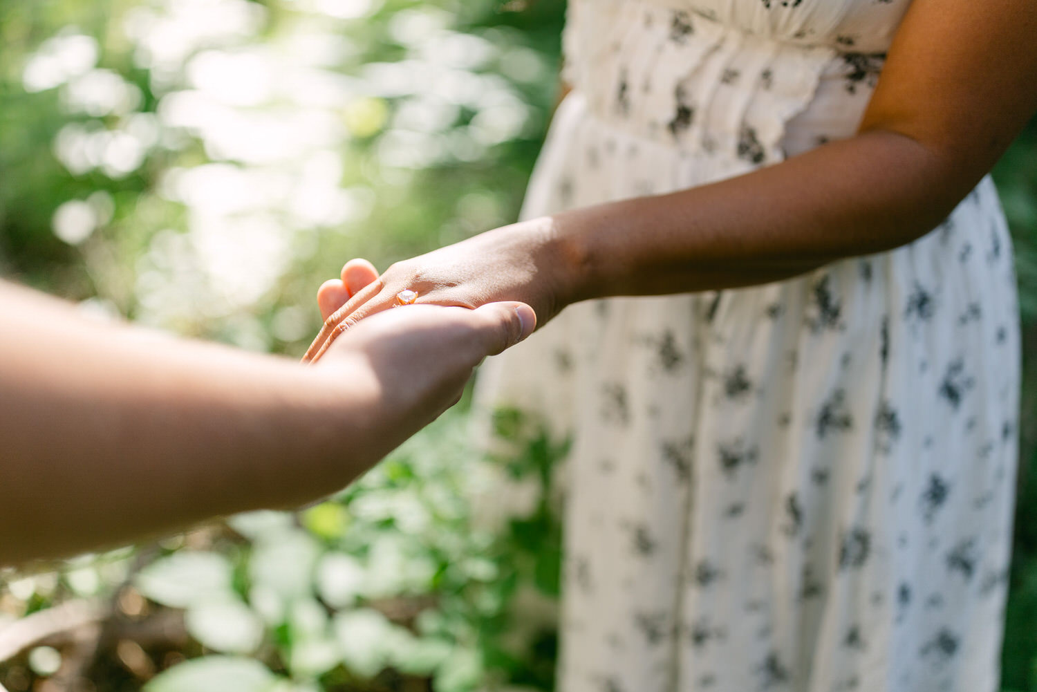 Two people holding hands, one wearing a floral dress, with a natural green backdrop.