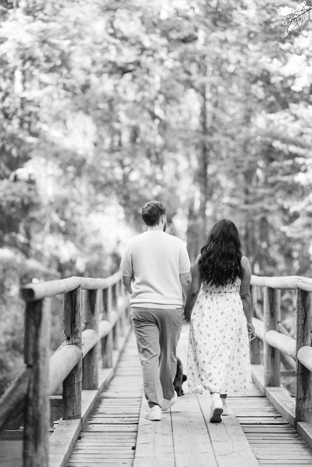 Black and white image of a couple holding hands and walking away on a wooden bridge surrounded by trees.