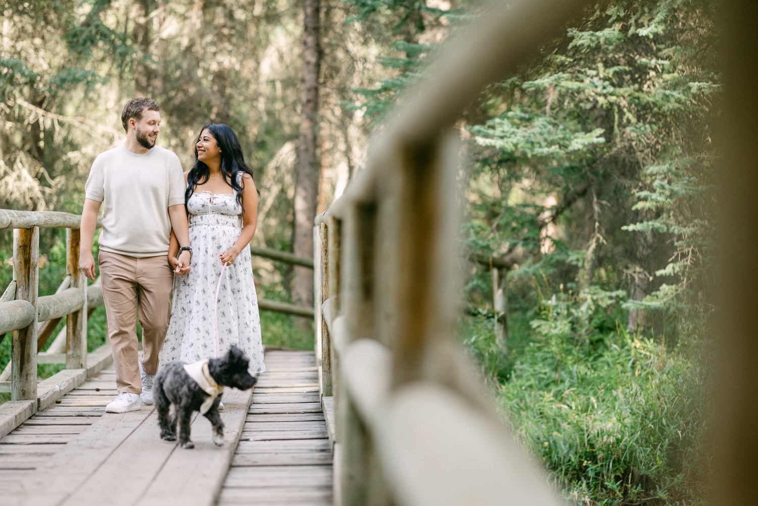 A couple holding hands and walking with their dog on a wooden bridge surrounded by lush greenery