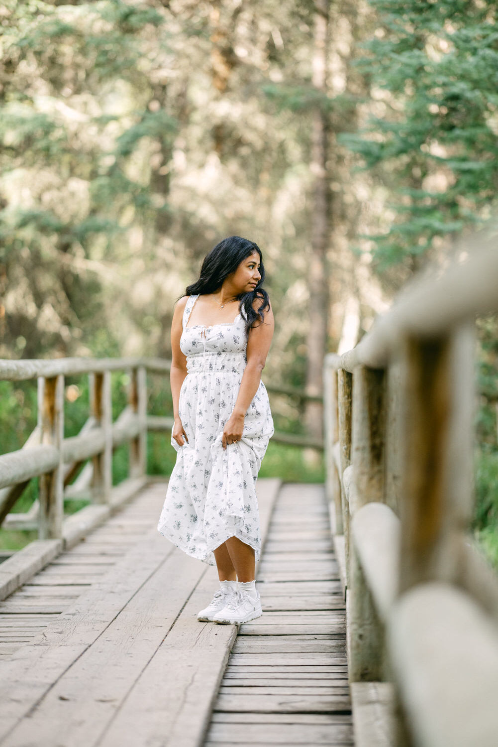 A woman in a white floral dress standing on a wooden bridge in a forest setting, looking to the side.