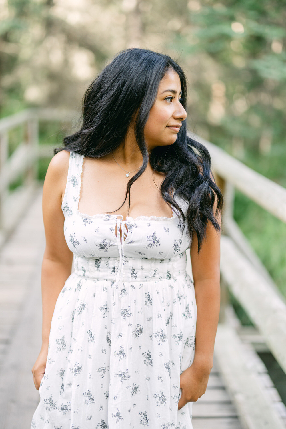 A woman in a white floral dress looking contemplatively to the side on a wooden bridge surrounded by green foliage.