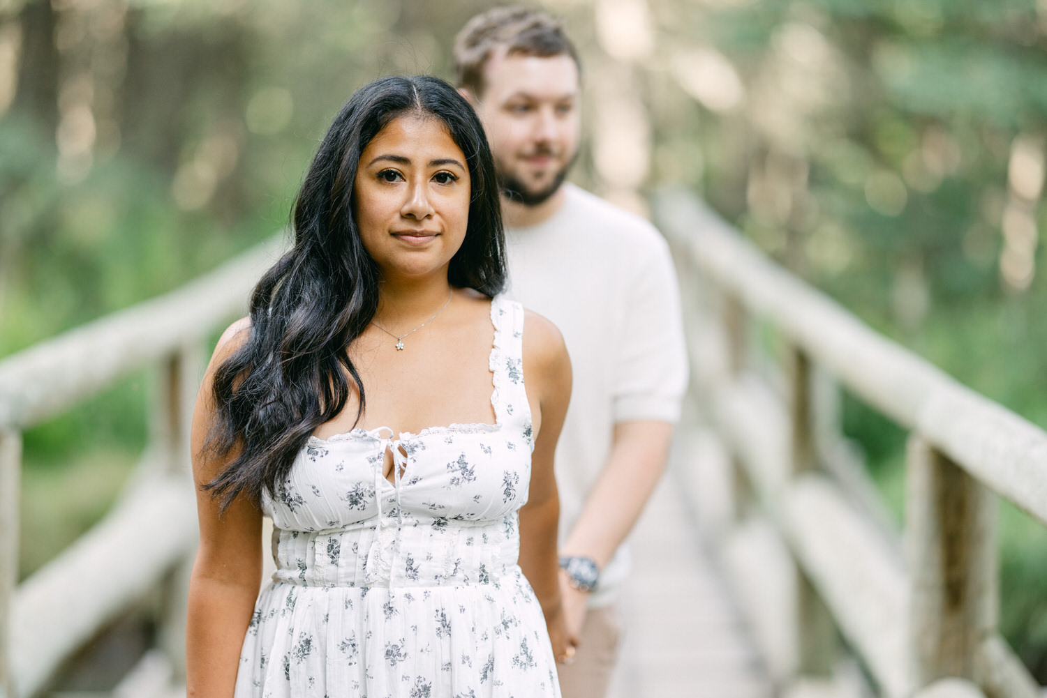 A woman in a floral dress standing in focus on a bridge with a man in the background.