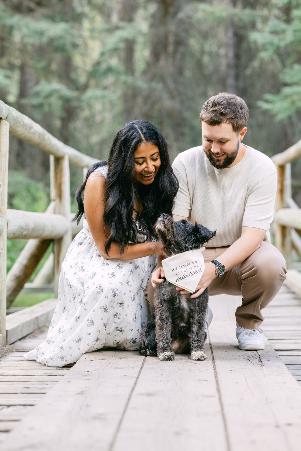 A happy couple crouched on a wooden bridge with their dog, which is wearing a sign saying "My humans are getting married."