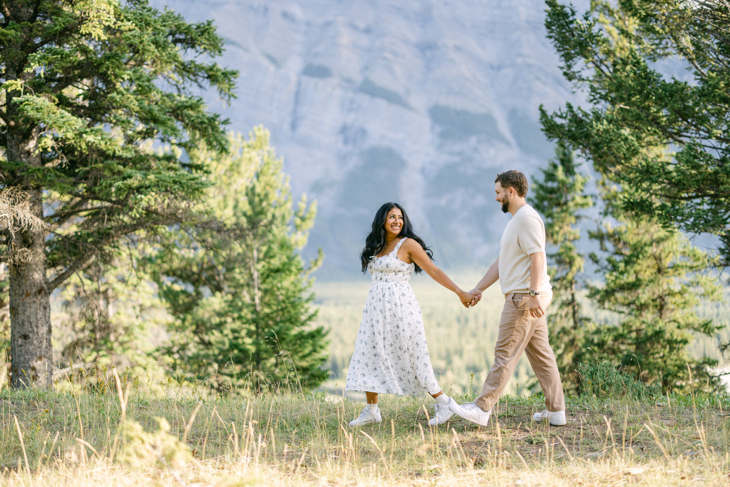 A man and a woman holding hands and walking through a grassy area with trees and mountains in the background.