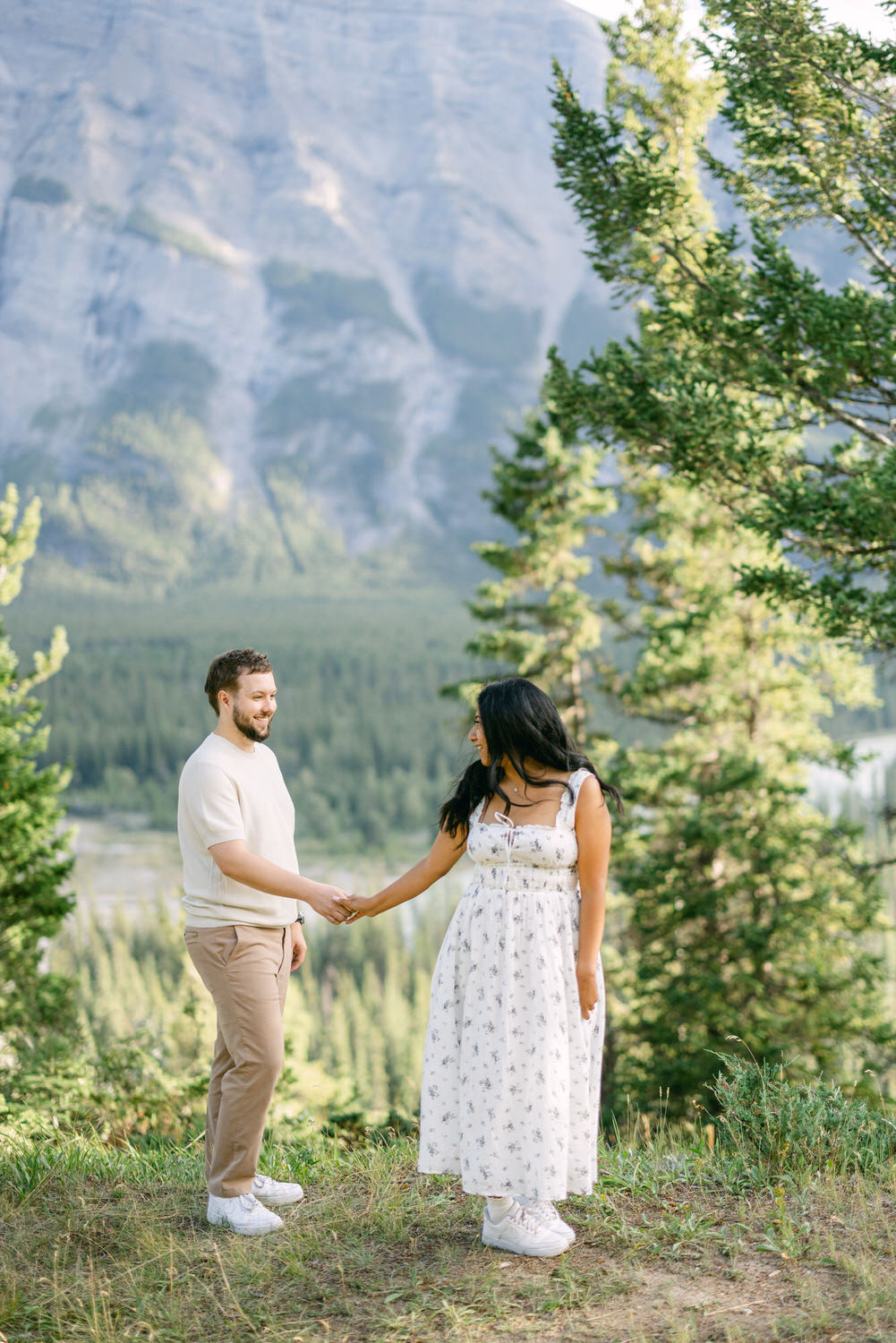 A couple holding hands in a serene mountain landscape, with the woman facing away and the man facing the camera.