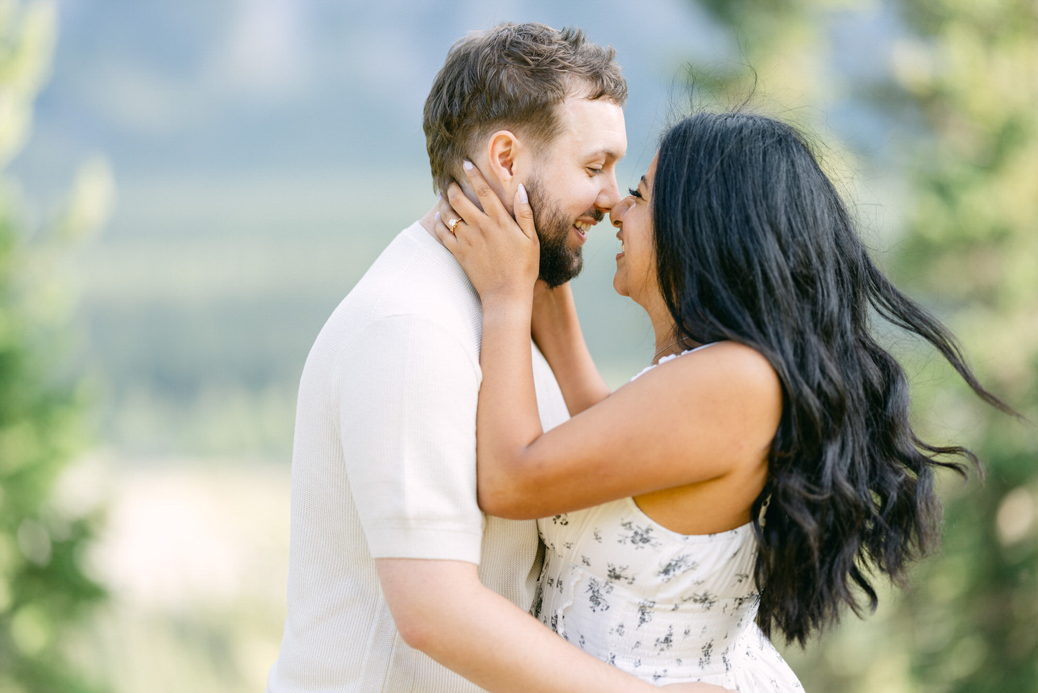 A man and woman affectionately touching foreheads and smiling while embracing outdoors with a blurred natural background