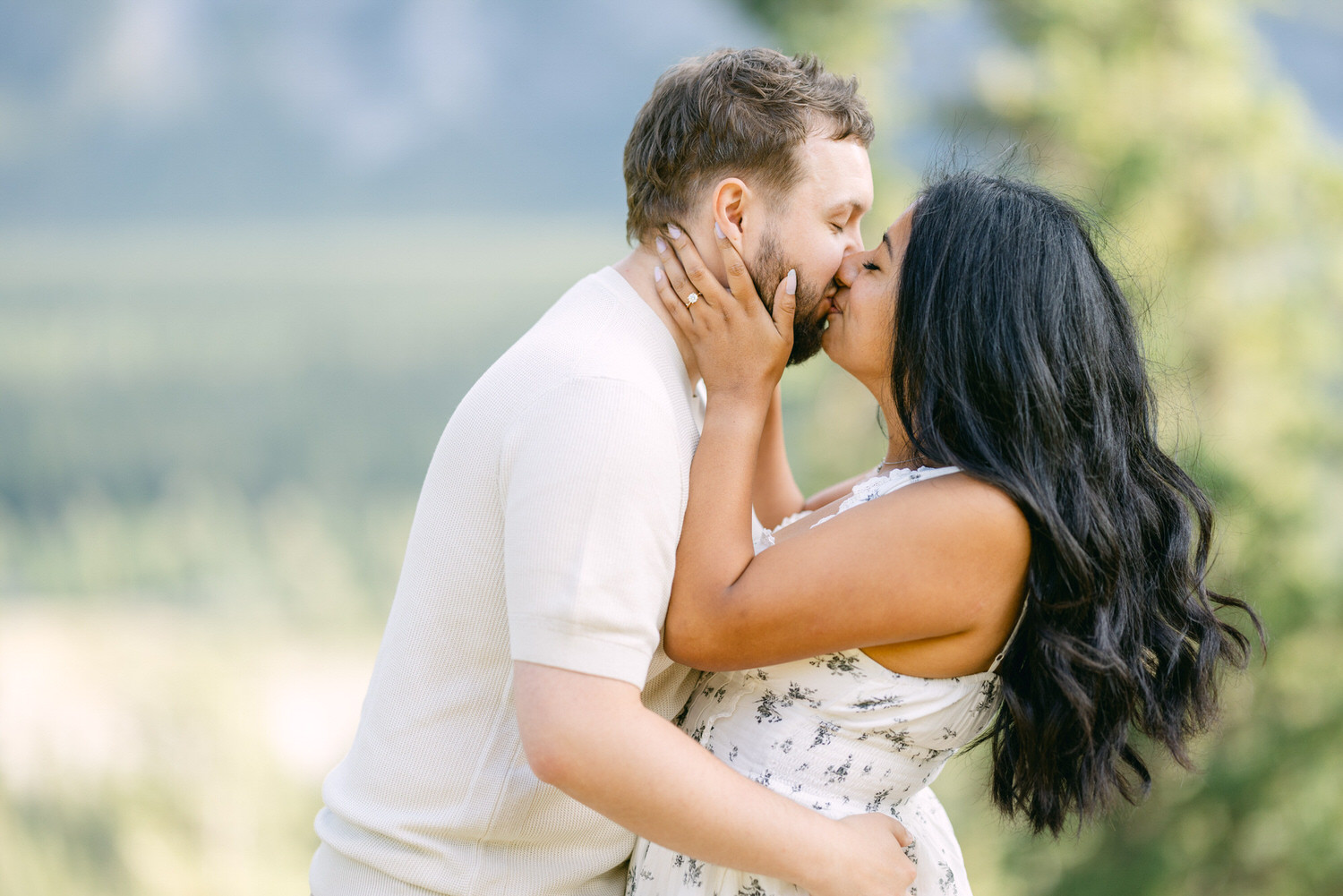 A man and woman sharing a romantic kiss outdoors with trees and nature in the background.