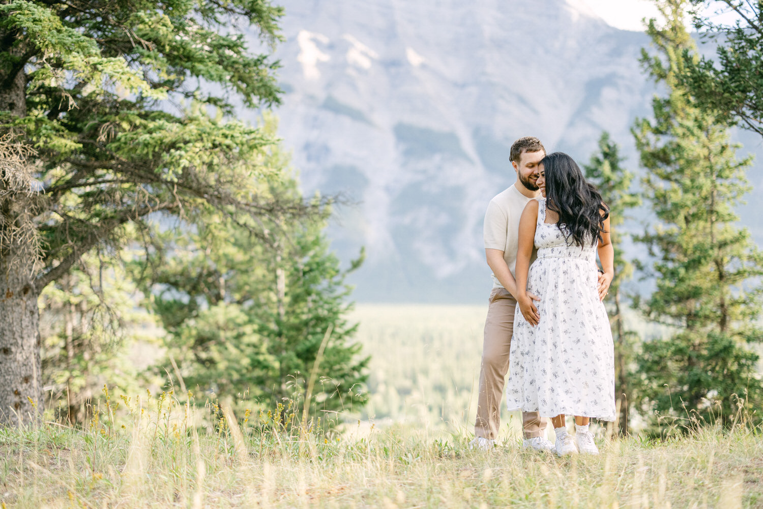 A couple standing closely together in a grassy meadow with trees and mountain backdrop, exhibiting affectionate body language.