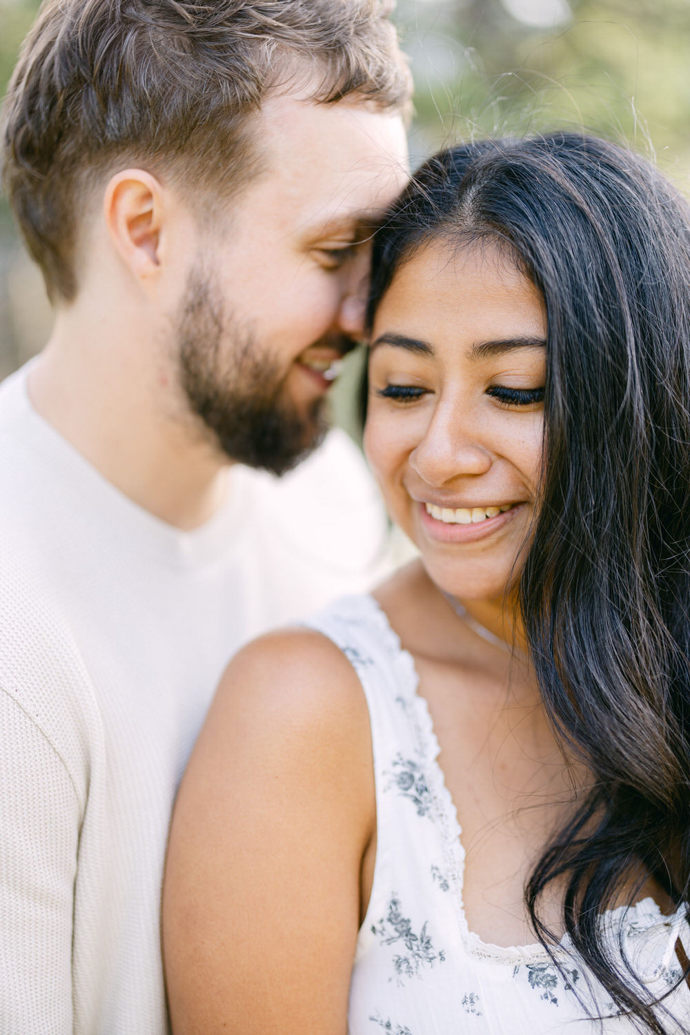 A close-up of a smiling woman with long black hair leaning on a bearded man in a white shirt, both looking happy and affectionate.