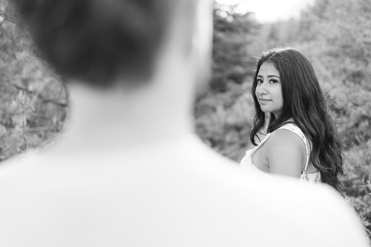 Black and white photo of a woman looking over her shoulder with another person's out-of-focus silhouette in the foreground.