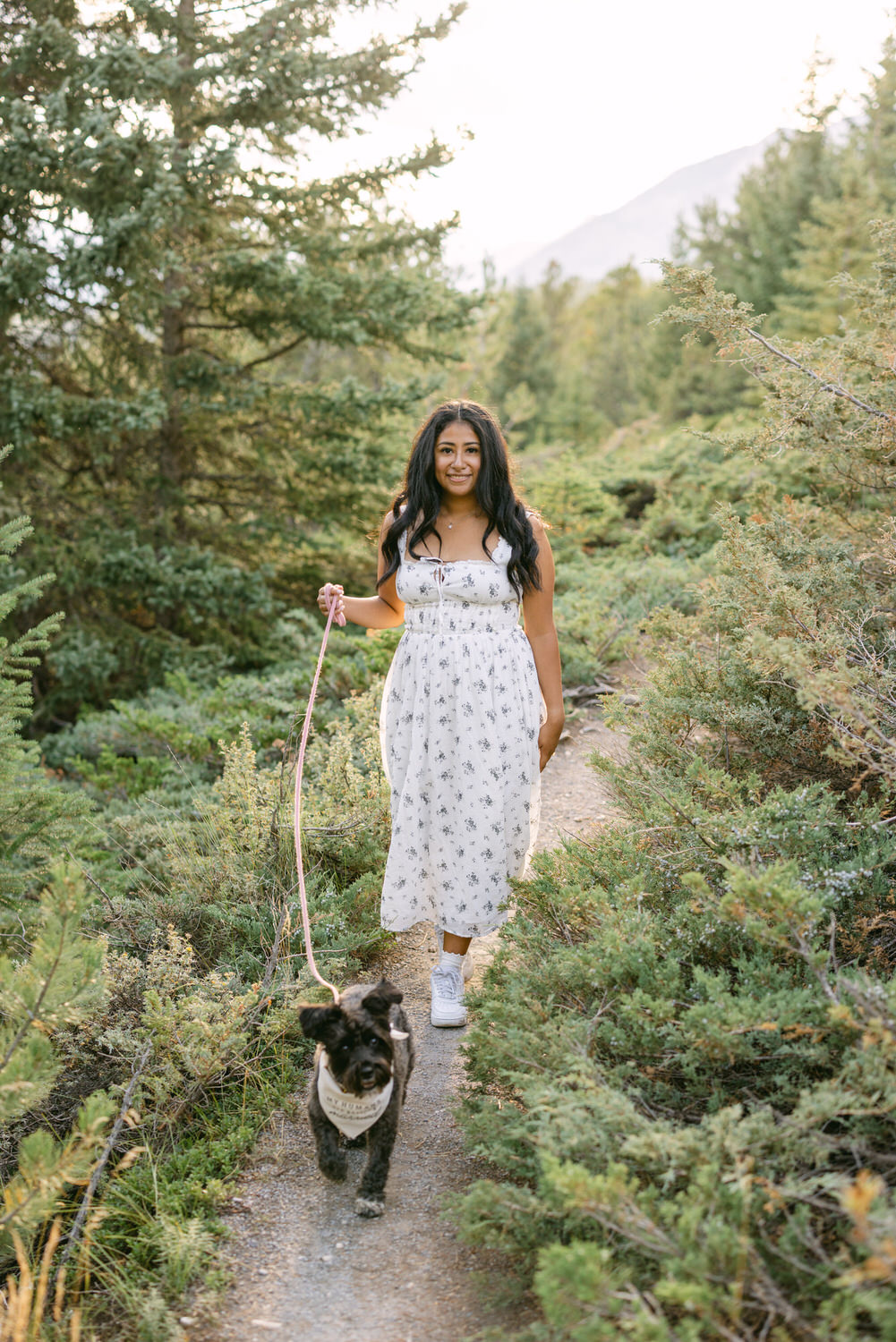 A woman in a white floral dress walking on a nature trail with her happy dog on a leash amidst greenery.