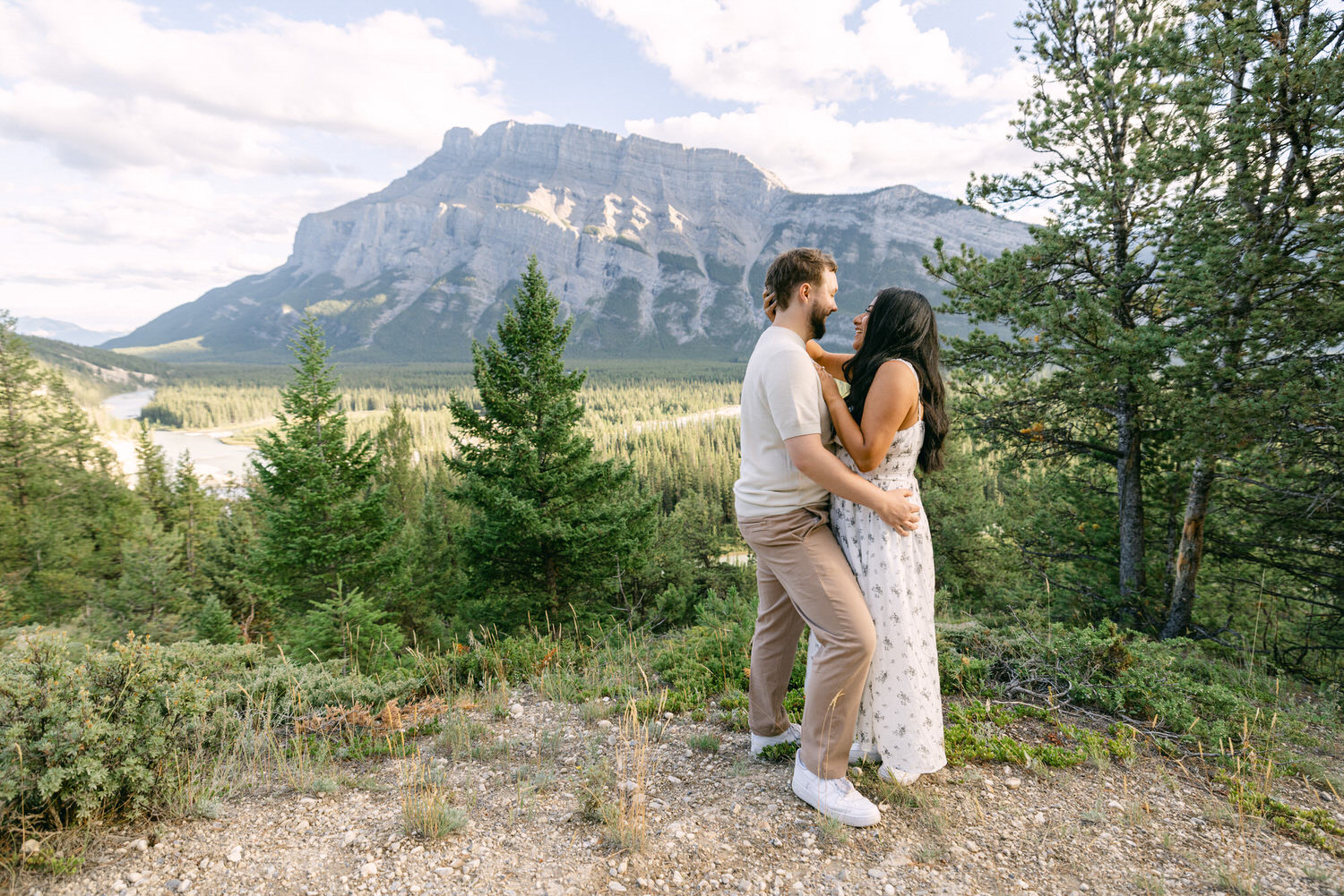 A couple hugging each other, with a serene mountain landscape in the background, featuring lush greenery and a calm river.