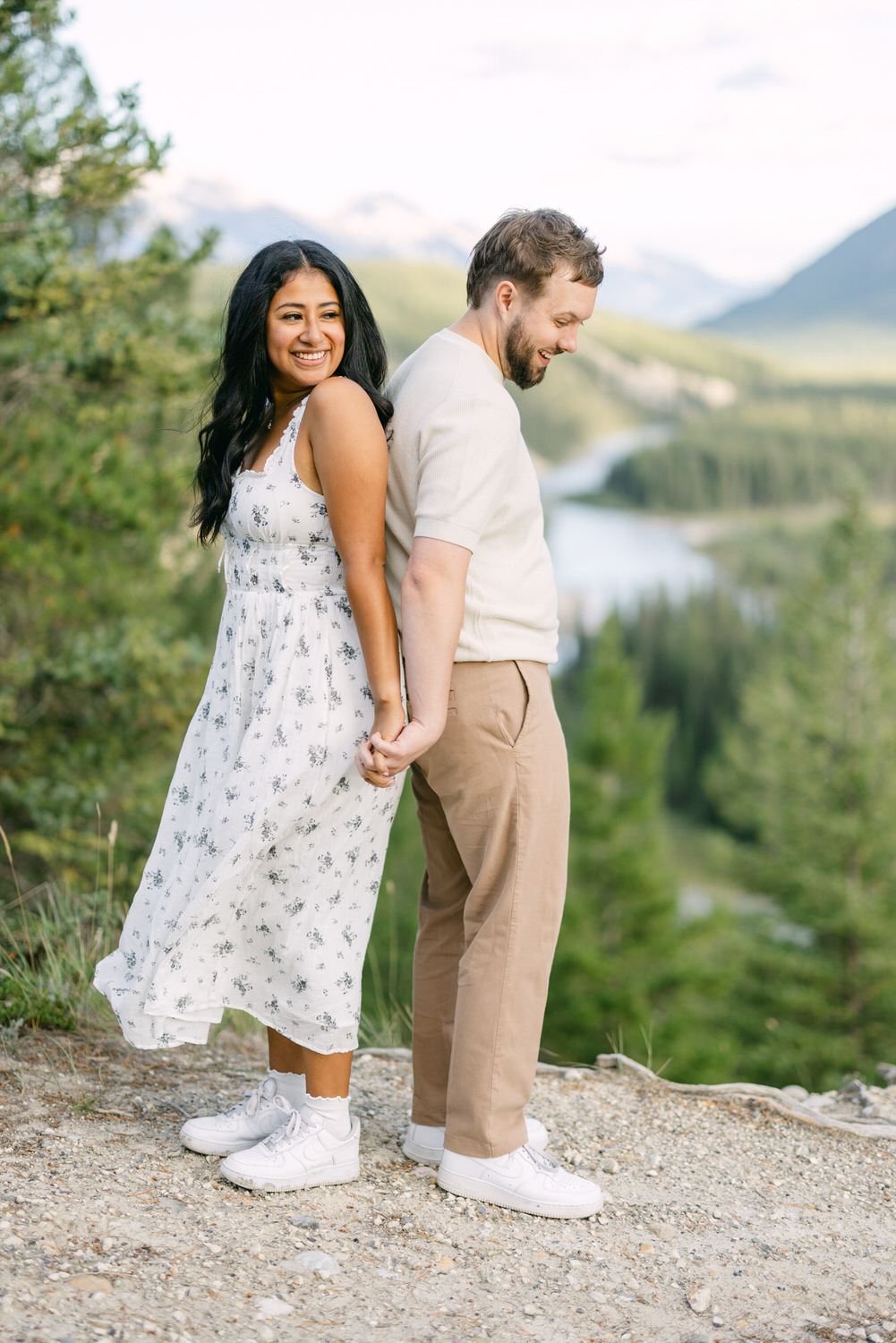 A man and woman holding hands and looking cheerful in a natural setting with mountains and a river in the background.