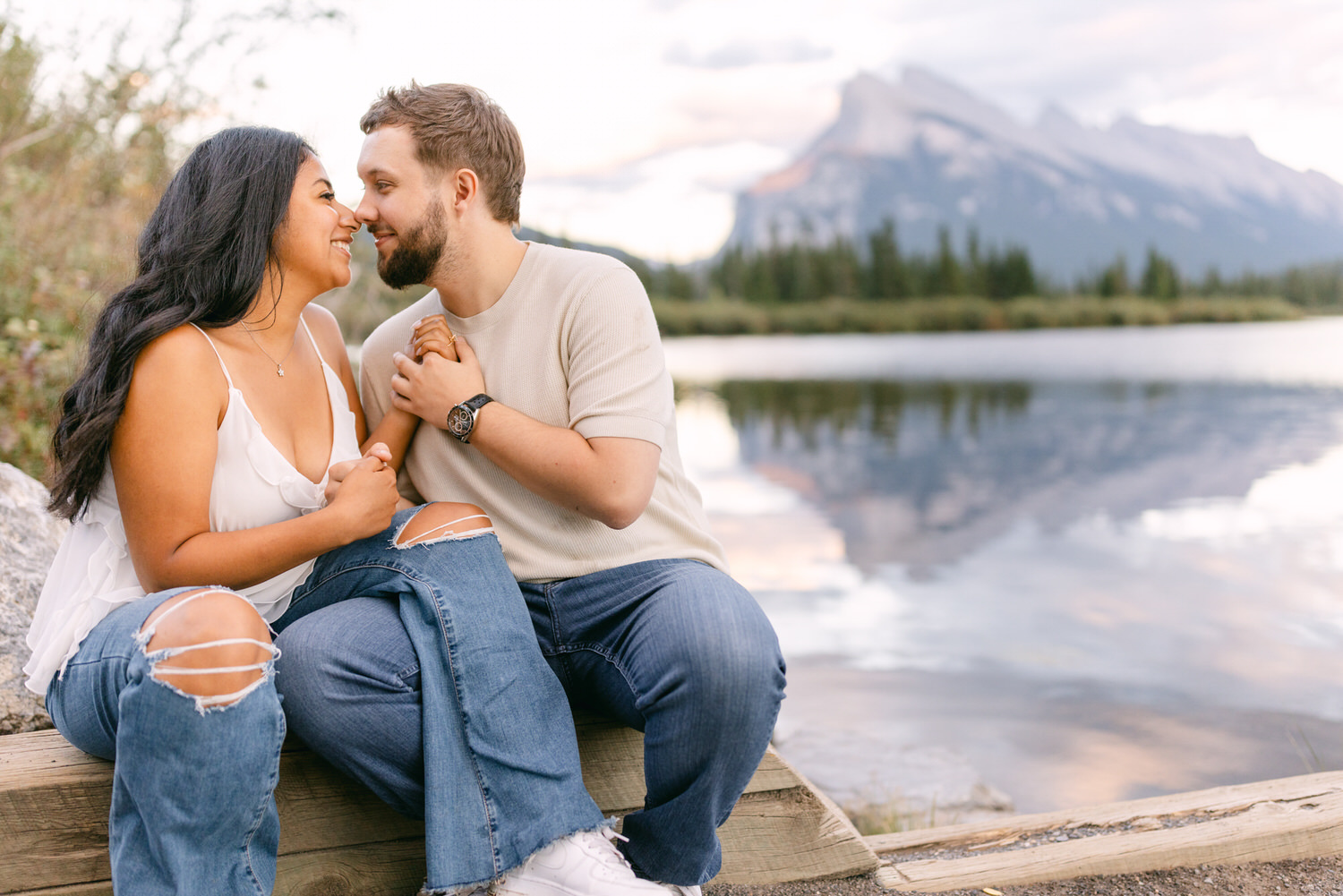 A couple sitting close to each other on a wooden dock, sharing an affectionate moment with a mountain and lake in the background.