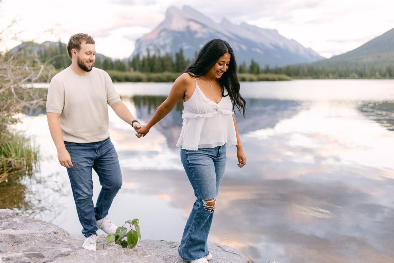 A man and a woman holding hands, walking along the shore of a serene lake with mountains in the background at dusk.