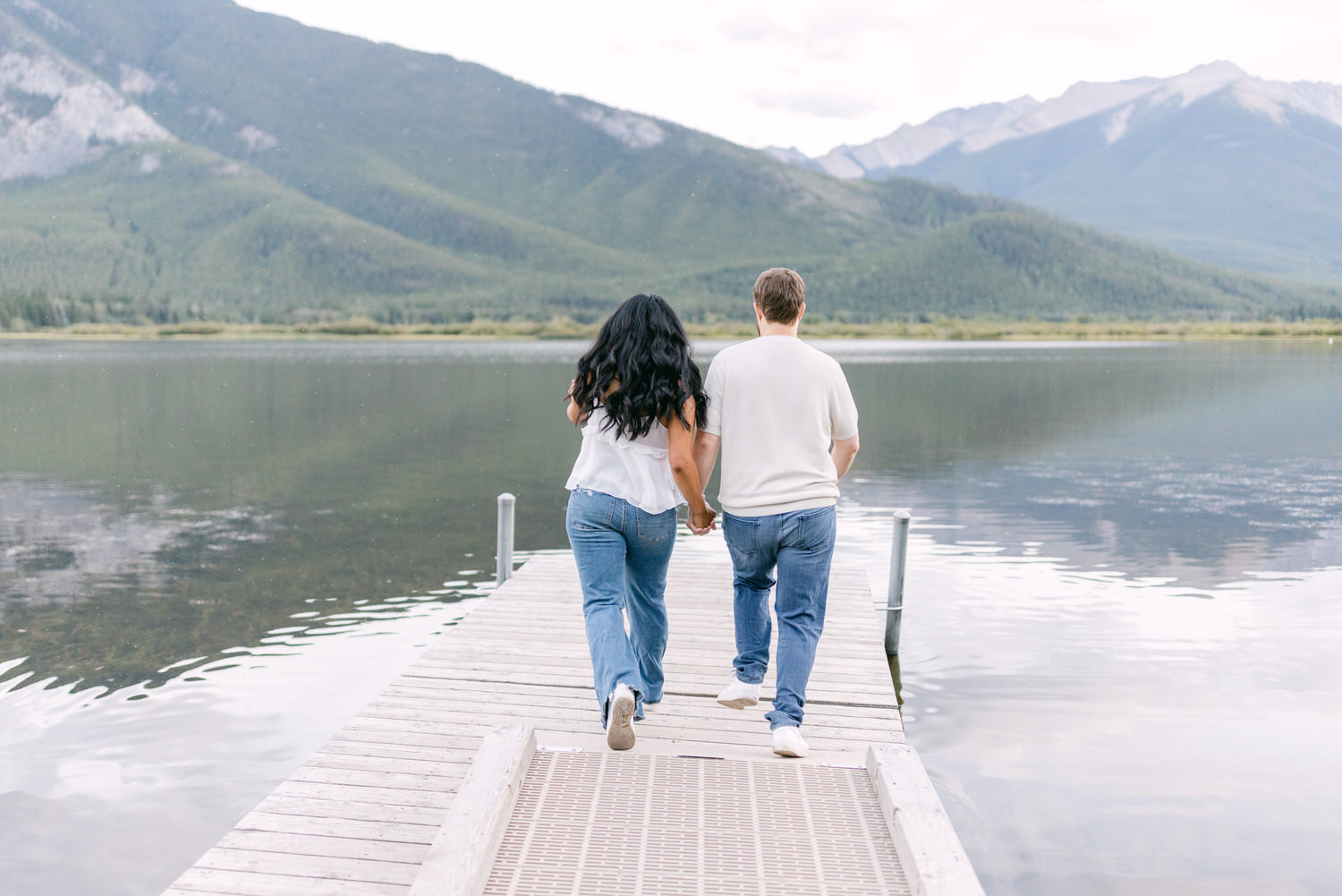 A couple holding hands and walking down a wooden pier by a tranquil lake with mountain scenery in the background.