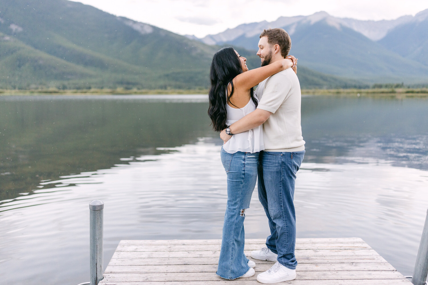 A couple hugging on a wooden dock with a serene mountain lake and forested mountains in the background.