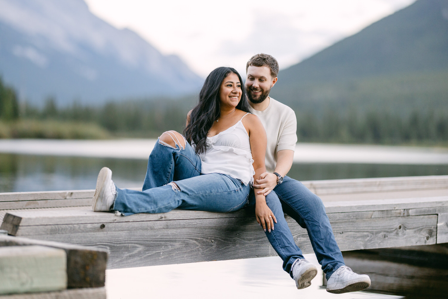 A man and woman sitting closely on a wooden dock with a serene lake and mountains in the background.