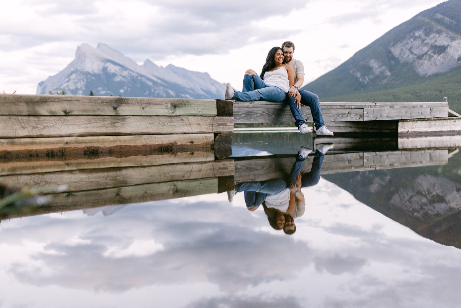 A couple sitting on a wooden dock with mountain reflections in the tranquil water.