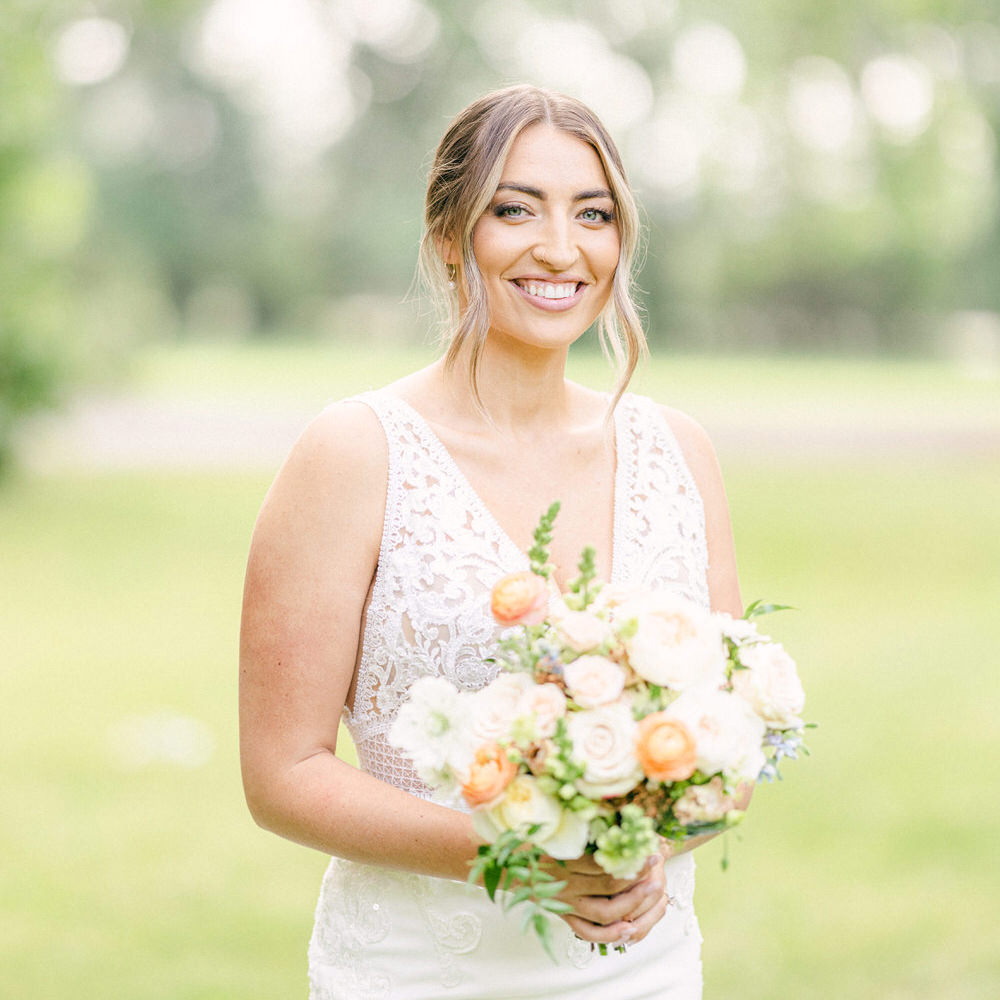 A joyful bride in a lace wedding dress smiling and holding a bouquet of flowers in a natural setting.