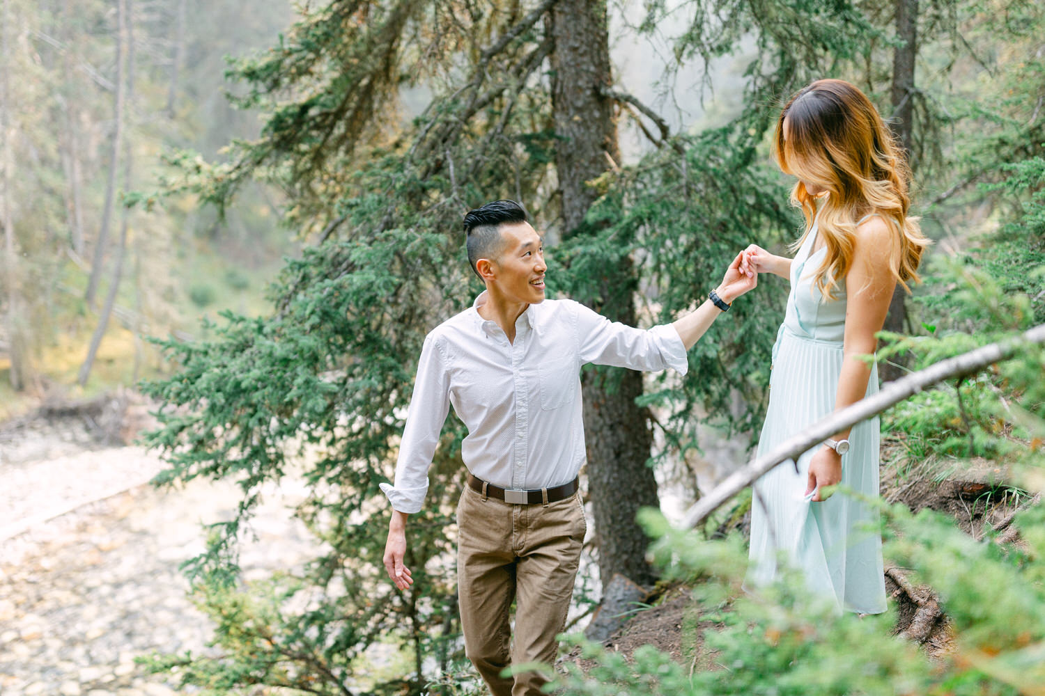 A man in a white shirt and beige pants holding hands with a woman in a pastel blue dress as they walk through a lush forest setting.