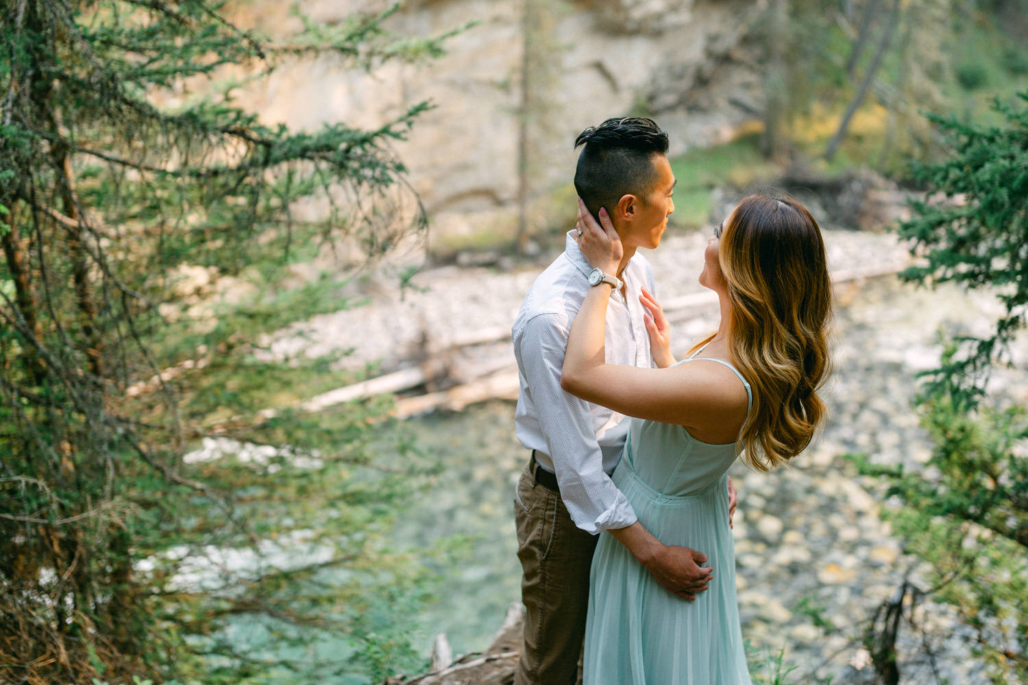 A couple shares a loving embrace in a serene forested area by a river.