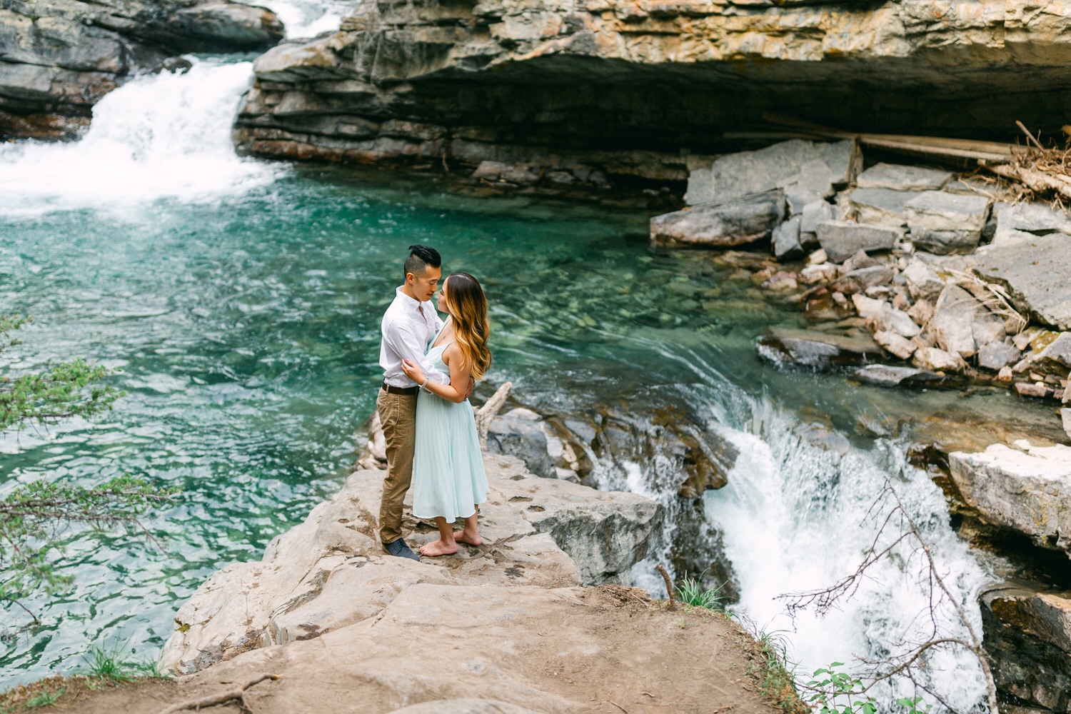 A couple hugging on a rock ledge in front of a small waterfall with clear blue water.