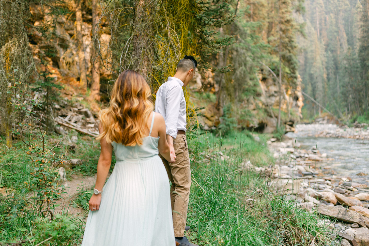 A man and a woman walking beside a river in a forested area, facing away from the camera.