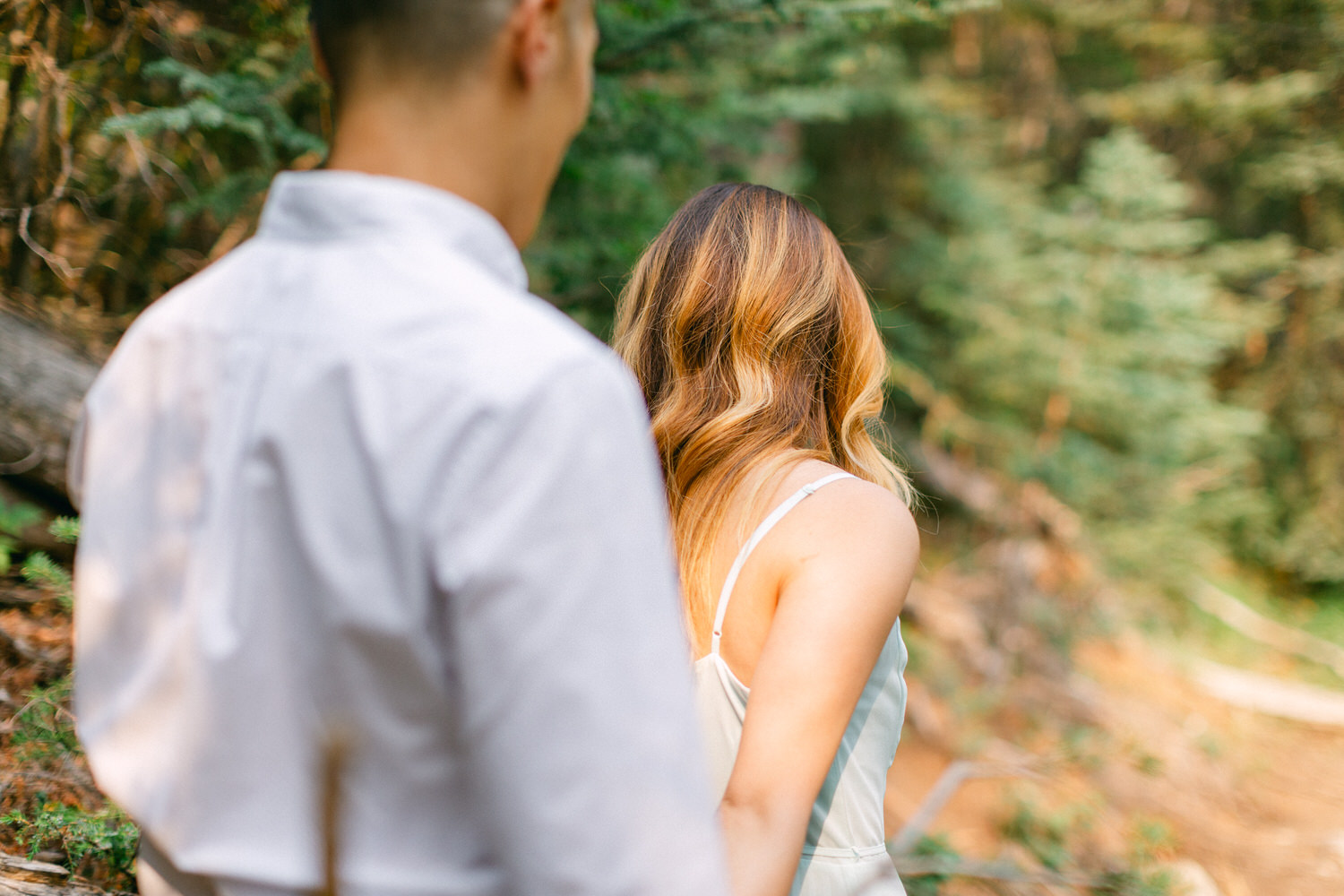 A man and a woman standing in a forest, woman facing away from the camera, man turned partially towards the camera with a blurred background.