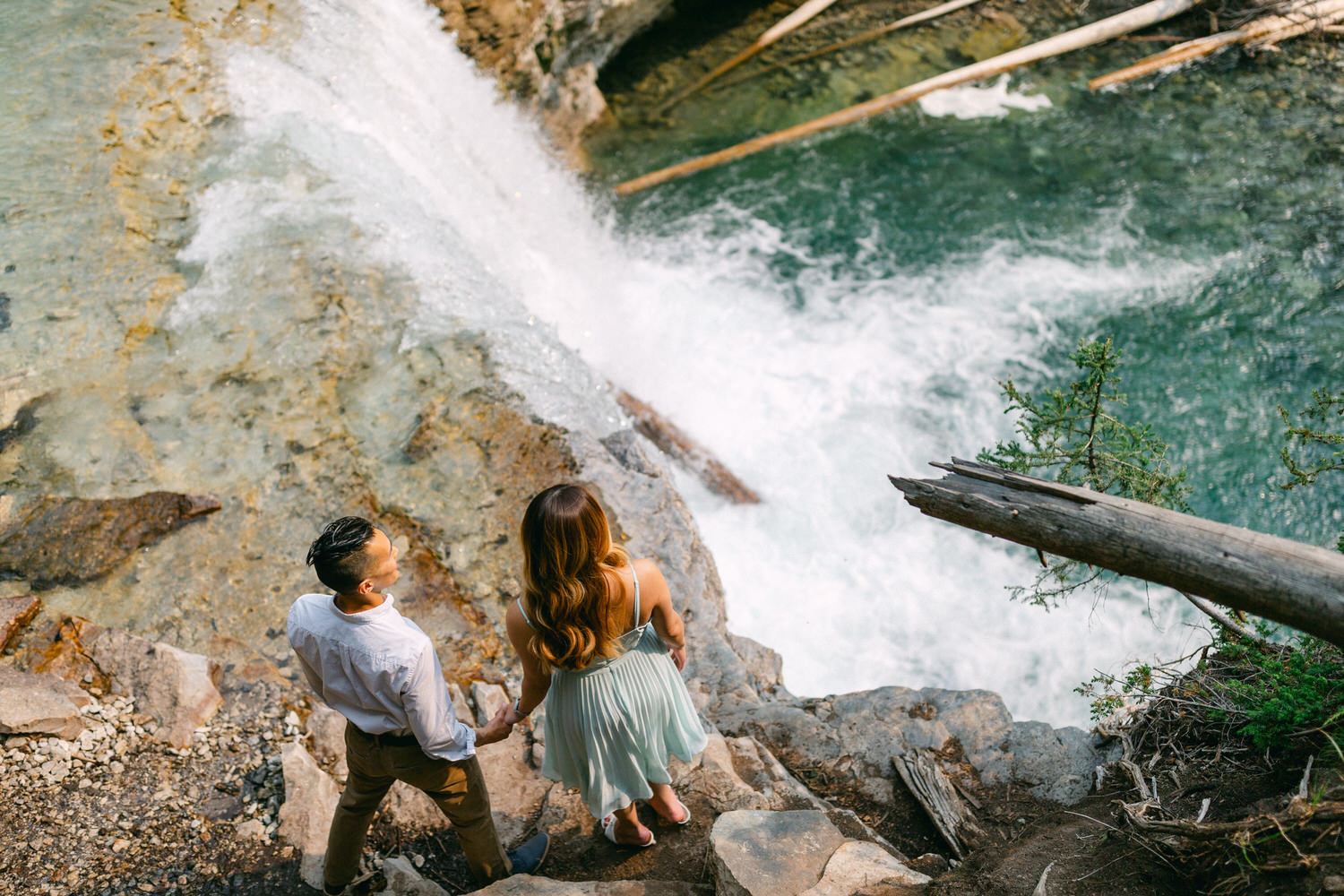 A couple holding hands and looking at a waterfall from a rocky vantage point.