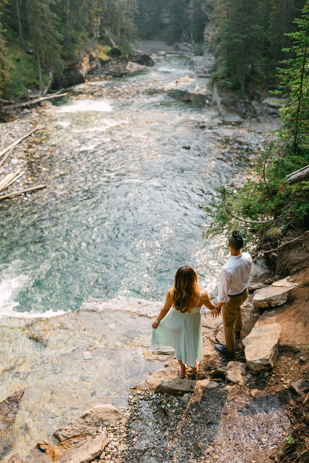 A couple holding hands while standing at the edge of a river with a forest in the background.