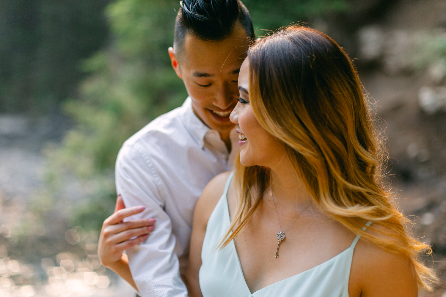 A man embracing a woman from behind, both smiling and enjoying a moment together outdoors with soft natural lighting.