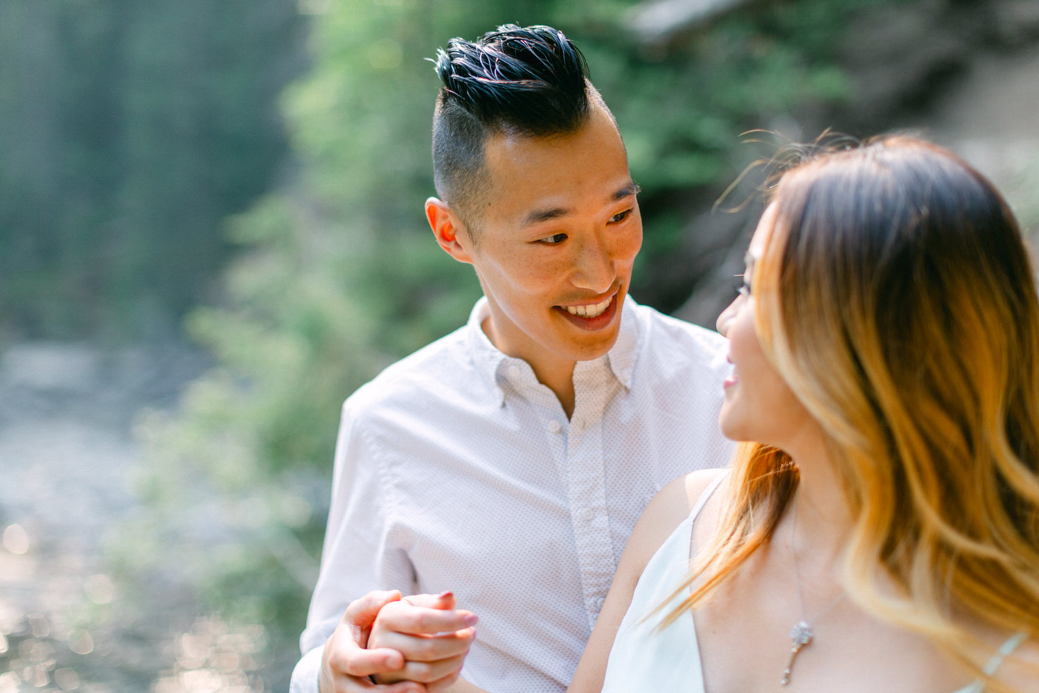 A smiling man holding hands with a woman, both dressed elegantly, with a blurred natural background.