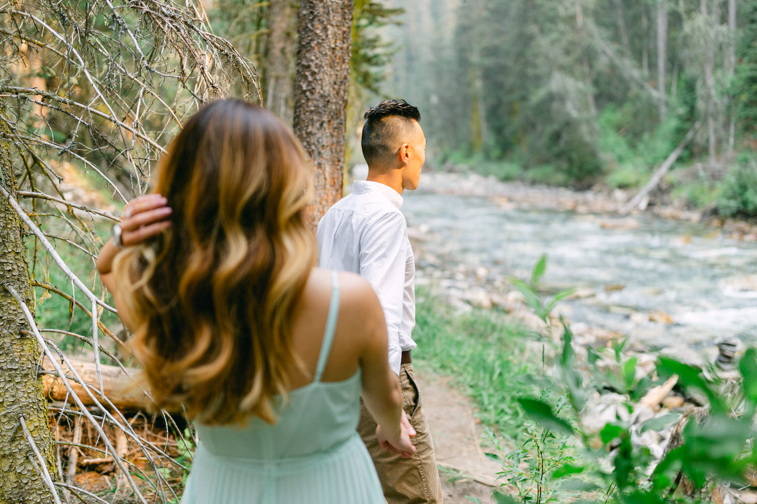 A person seen from behind looking out over a river, with another person partially visible in the foreground, set against a backdrop of lush greenery and flowing water.