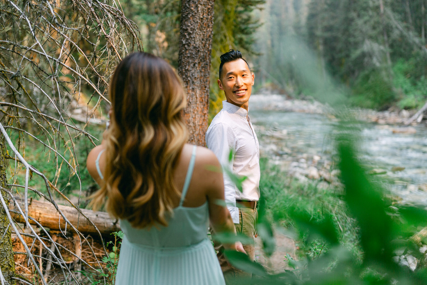 A man smiling at the camera while a woman with her back turned looks on, both standing by a forest river.