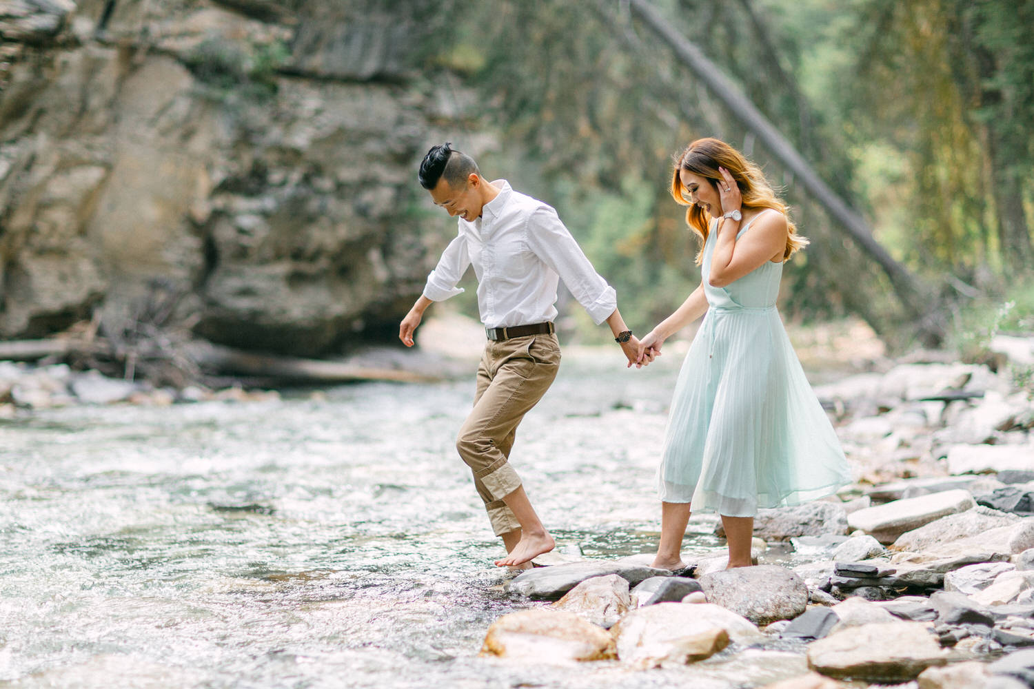 A man and a woman holding hands while carefully walking on rocks across a river.