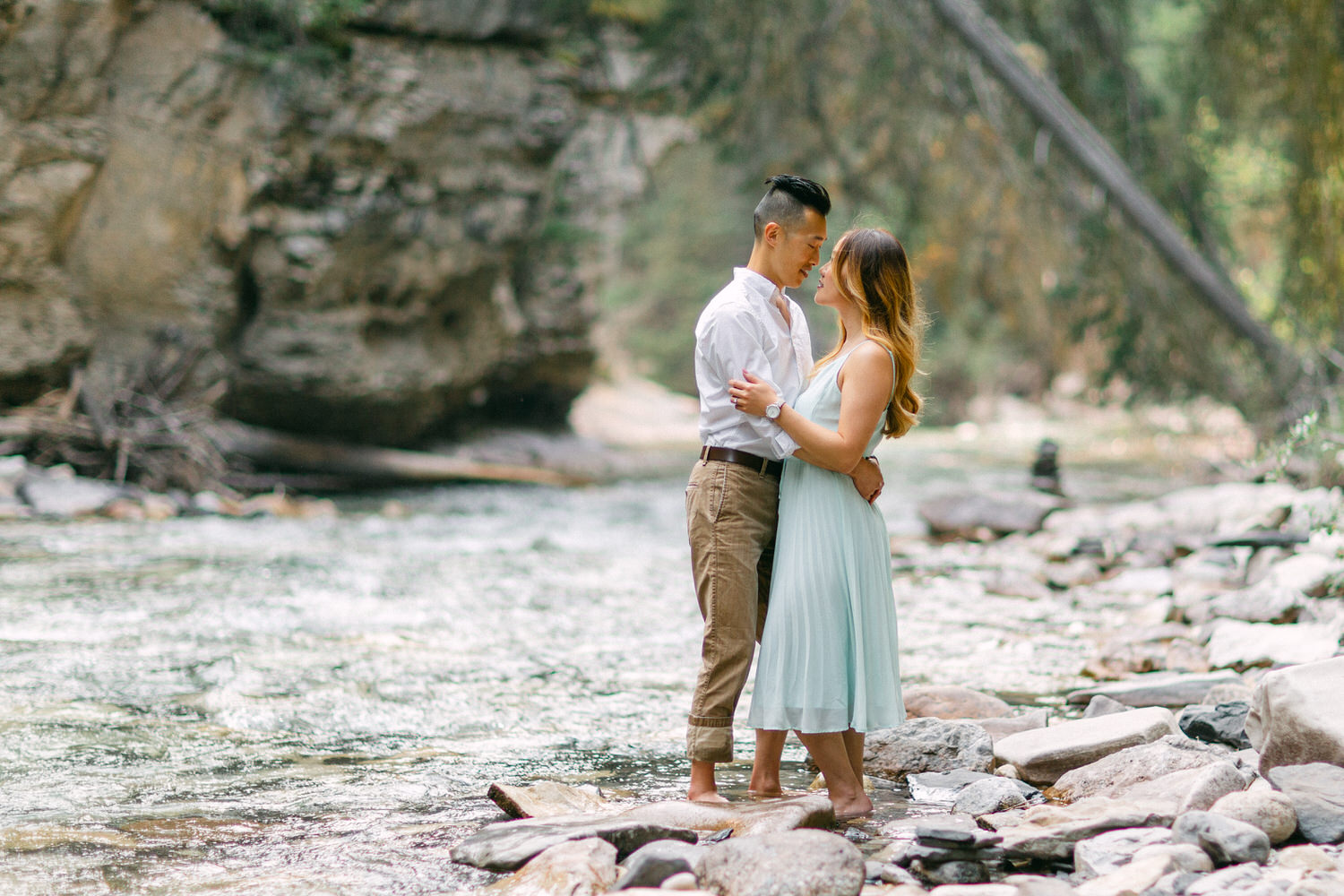 A couple holding each other while standing on rocks beside a gently flowing river, surrounded by a forested area.