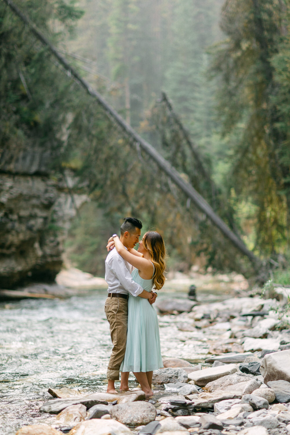 A couple holding each other in a loving embrace by a rocky river bank, surrounded by forest.