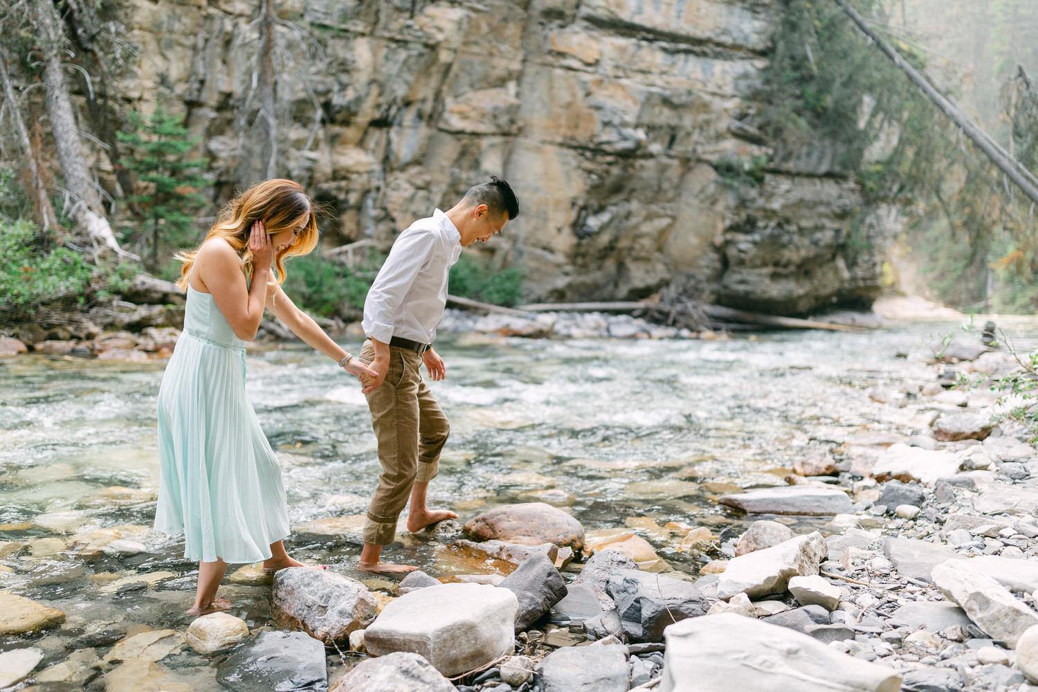 A man and woman holding hands while carefully stepping on rocks to cross a river, with forest and rocky terrain in the background.