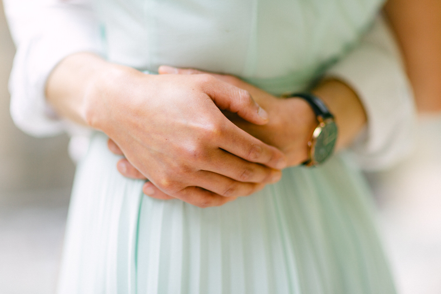 A person's hands gently clasped together wearing a watch with a focus on a pale blue dress in the background.