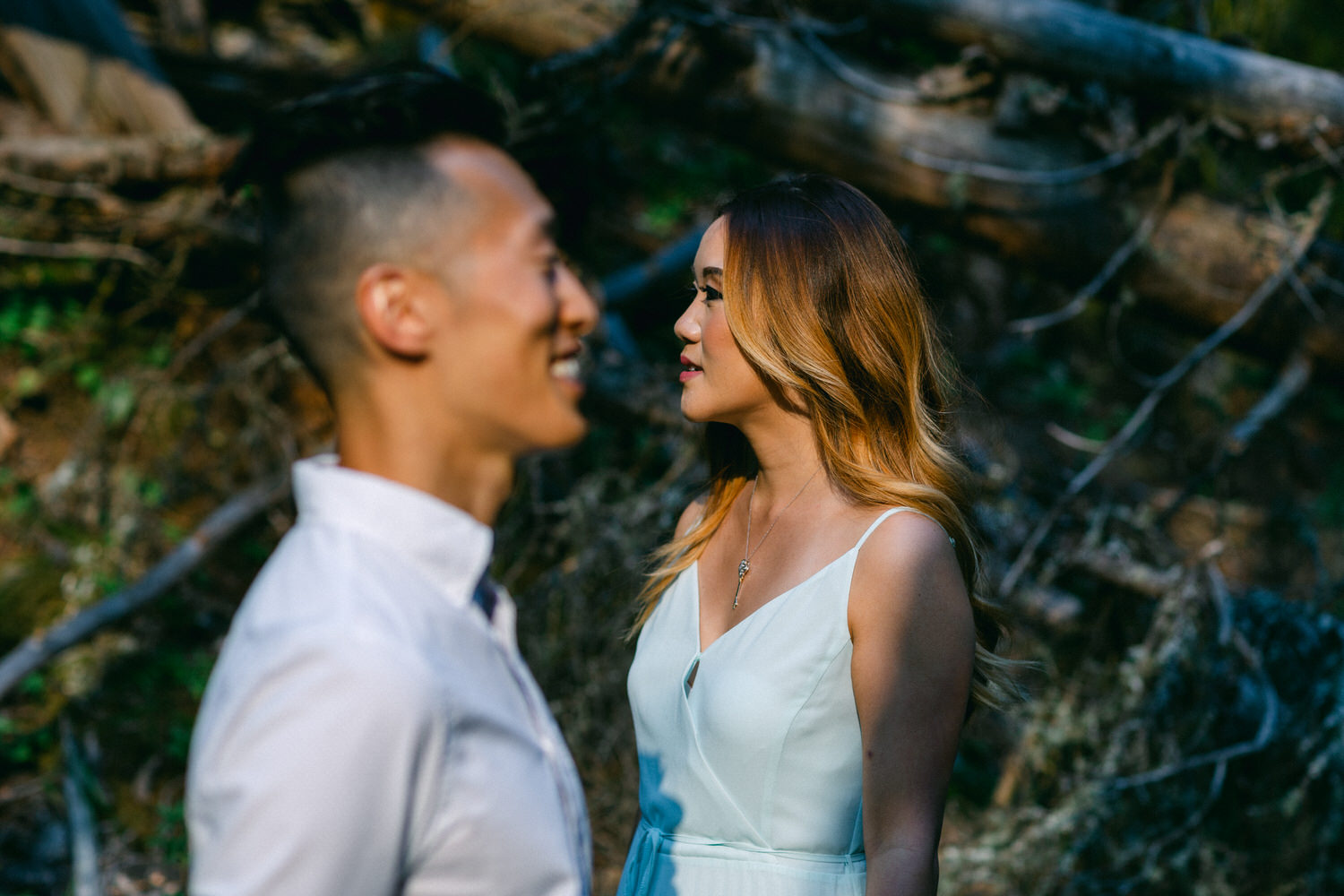 A man and a woman smiling at each other in a forest setting with sunlight filtering through the trees.