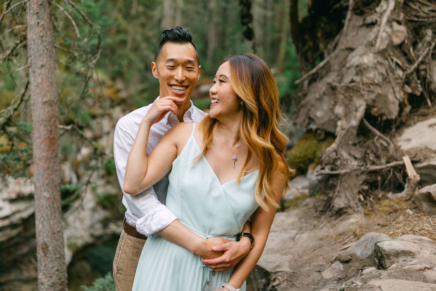 A man and woman smiling and embracing each other in a wooded area with trees and a fallen tree trunk in the background.