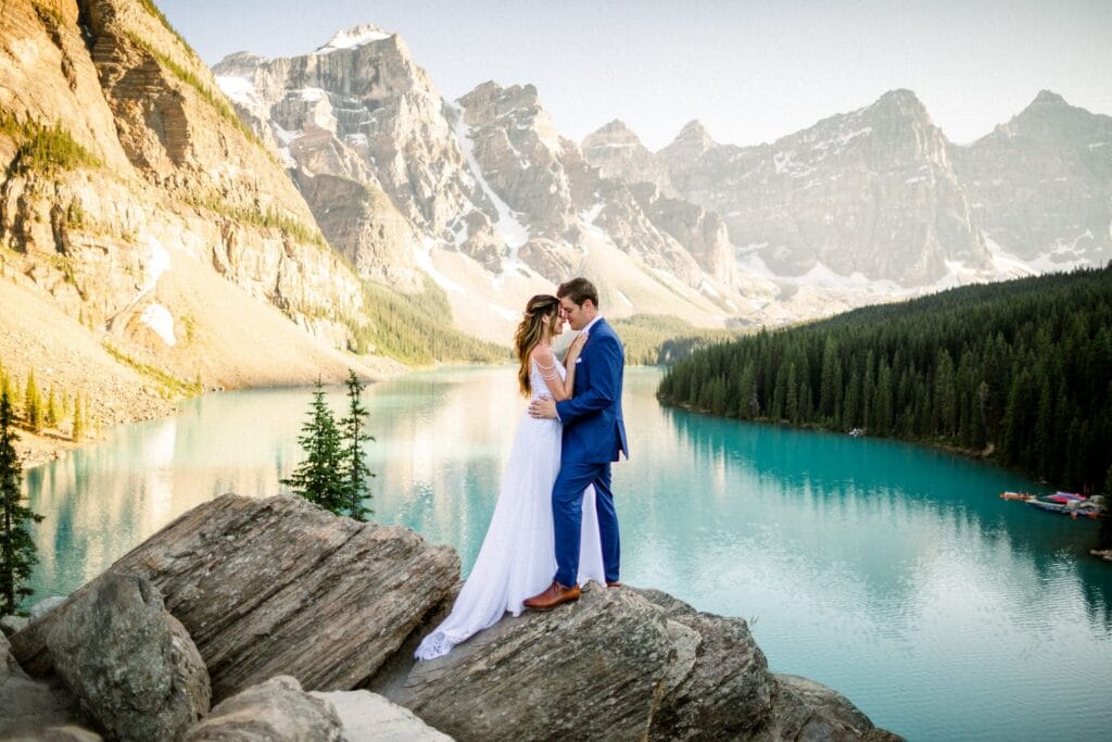 A couple sharing a kiss on a rocky lakeside with mountains and a turquoise lake in the background.