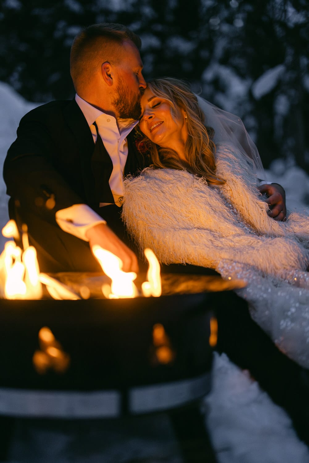 A bride and groom sharing an intimate moment by a fire pit in a snowy setting.