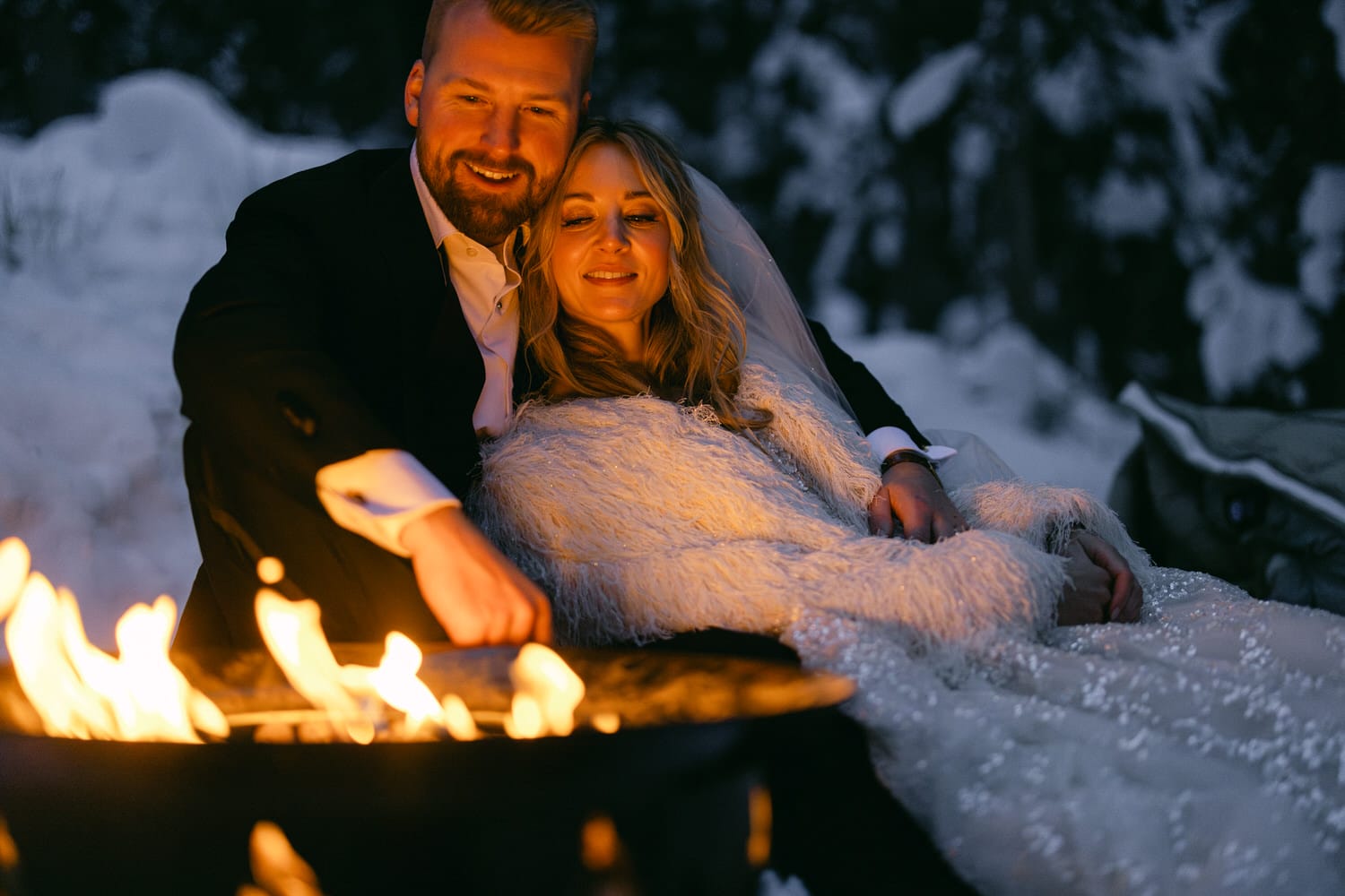 A couple in wedding attire cuddling by a fireside in a snowy setting at dusk