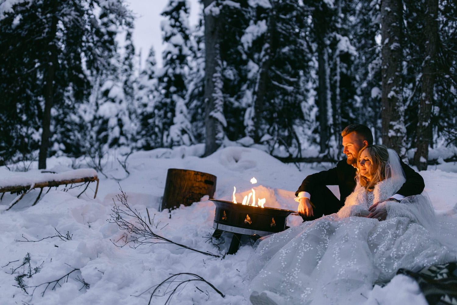 A couple in wedding attire cuddling by a small campfire in a snowy forest setting.